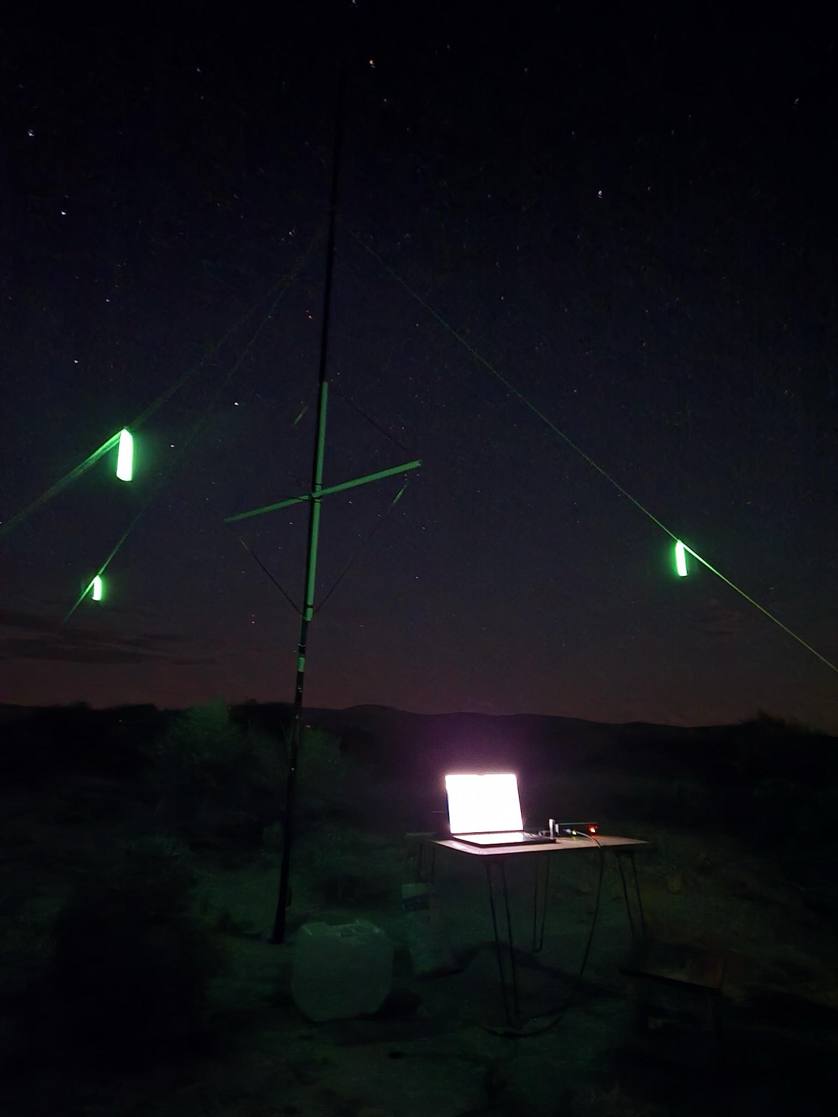 A night field station under a starry sky: a tall mast with guy lines marked by green glow sticks, and a laptop lit up on a folding table in the scrub.