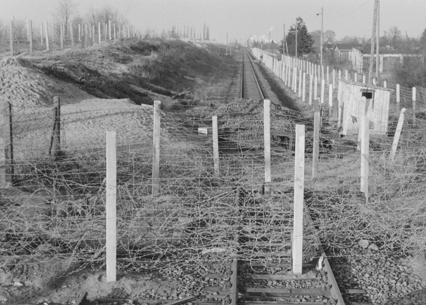A black and white photograph of a railway line running through a border strip, with rows of concrete posts and coils of barbed wire strung across the tracks.