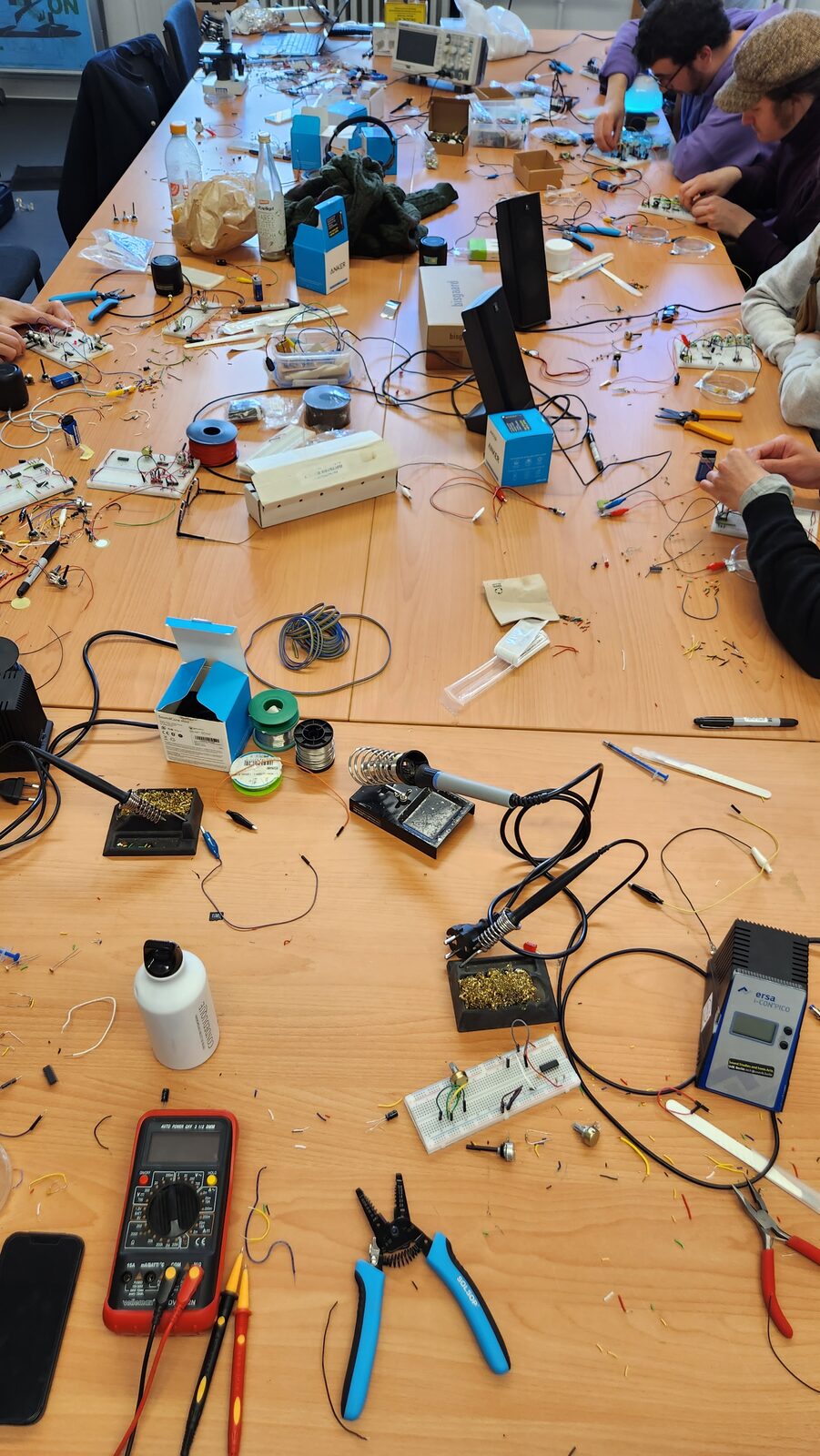 A workshop bench covered with soldering stations, breadboards, components and tools, with participants building circuits.