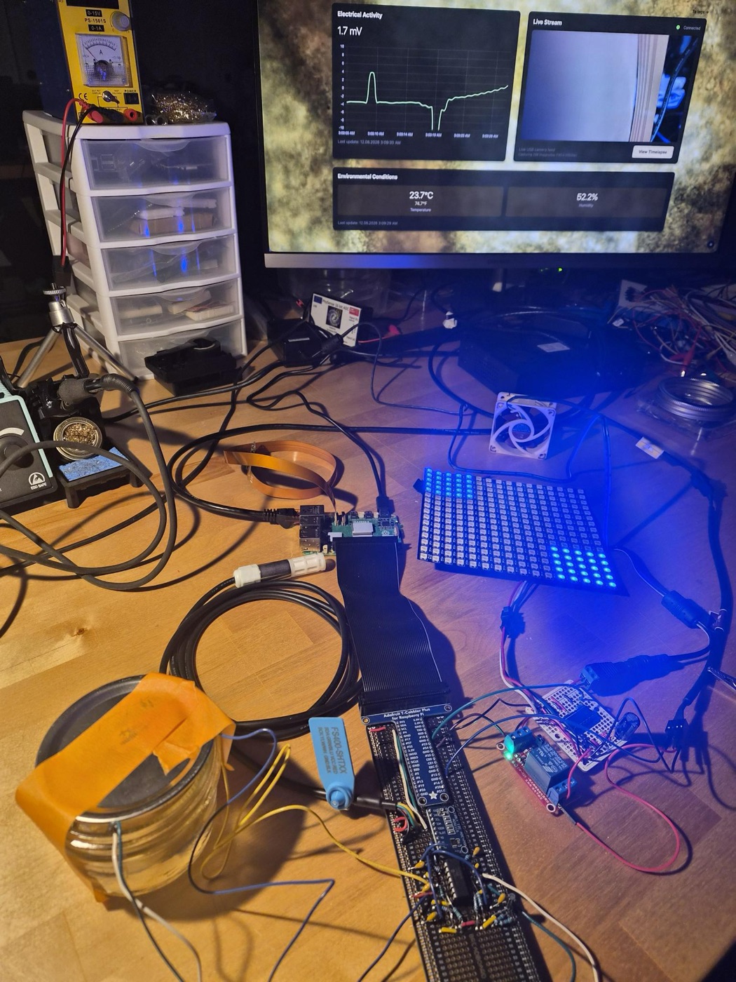 A workbench with an LED matrix glowing blue, a Raspberry Pi, a humidity sensor, a jar wrapped in tape, and a monitor showing an electrical activity graph and live stream.