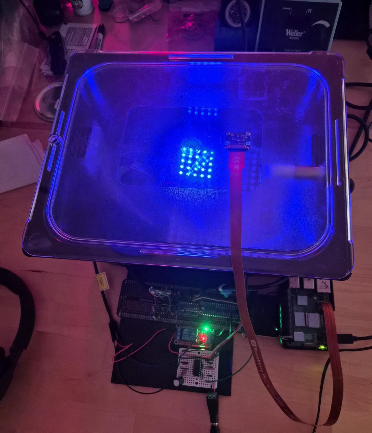 A sealed transparent chamber lit from below in blue, with a cluster of LEDs glowing under the lid and a camera module mounted on top.